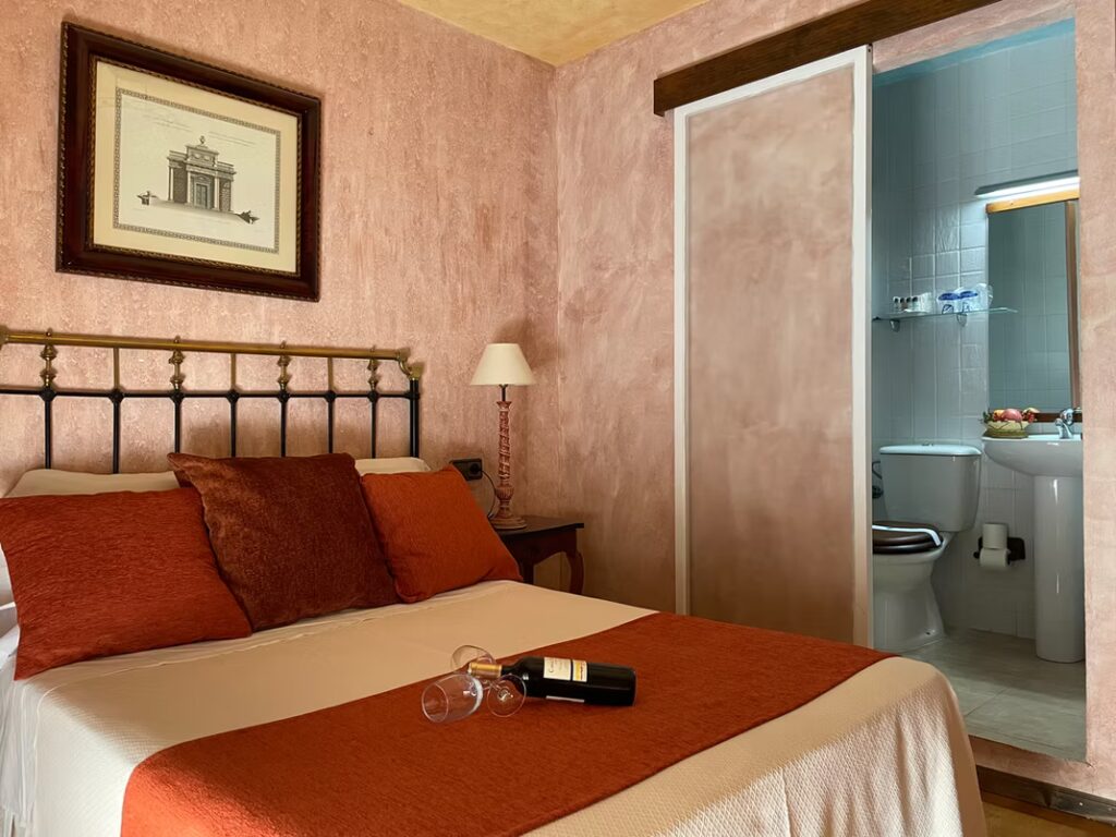 Habitación doble de la Posada del Hidalgo con cama de matrimonio, colcha y cojines color caldera, mesillas de madera y acceso a baño privado con lavabo y espejo iluminado.